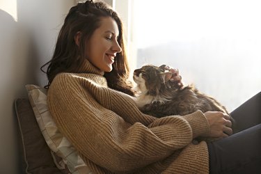 A young woman by a window is looking happily at a cat that is sitting in her lap.