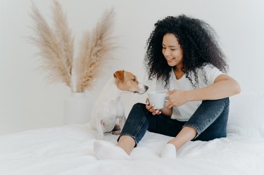 Happy brown-skinned woman with dog and coffee mug on the bed