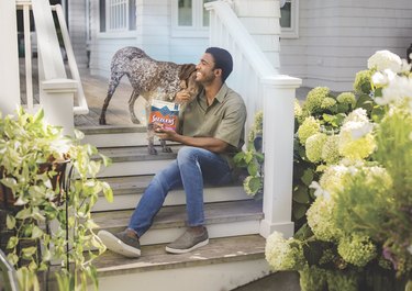 man sitting on steps holds bag of Blue Buffalo treats and pets dog